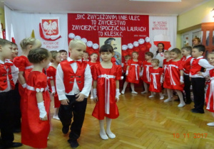Na tle biało czerwonej dekoracji stoją w półkolu dzieci ubrane na biało-czerwono.Jedna para stoi na środku.